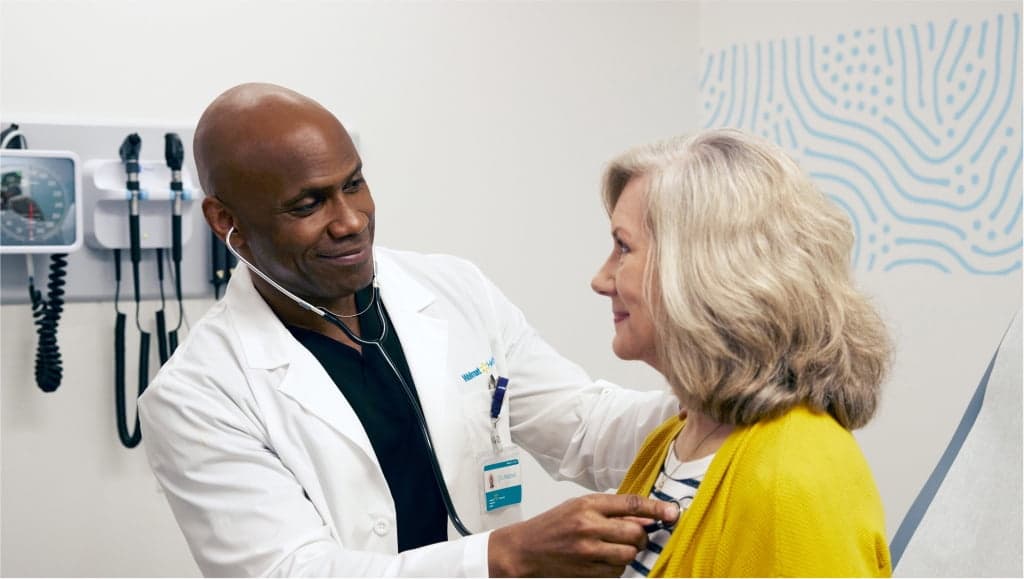 Image of a male doctor using a stethoscope on an older female patient at Walmart Health 